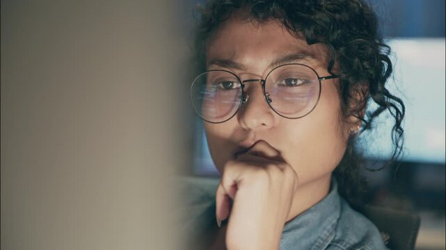 Close-up of a young female developer with glasses reflecting code, deeply thinking during late-night work session in a dimly-lit workspace.