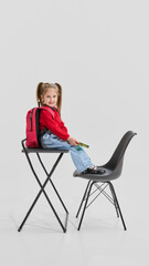 Fototapeta premium Little girl with pigtails wearing red sweater and jeans sitting on school desk with backpack, smiling confidently. Concept of first day, primary education, self-expression and new learning goals.