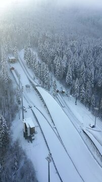 Aerial view of snow-covered ski jumping hill surrounded by dense winter forest on a cold, foggy day with soft light. Concepts of winter sports, nature, and travel.