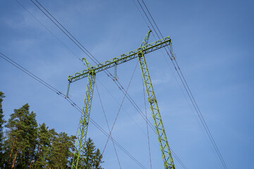 Fototapeta premium High Voltage Power Line Tower in Forest Area. Tall green metal power line tower with high voltage wires stretching across a blue sky, standing near a forested landscape.
