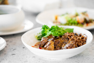 A close-up of a white bowl filled with flavorful chicken, topped with sauce, served with brown rice and fresh greens, against a softly blurred restaurant background.