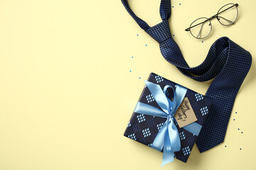 Father's Day flat lay: gift box, blue necktie, glasses, and confetti on yellow background. Top view with copy space.