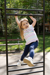 Happy little girl hanging on the horizontal bar on a sunny day. Children's fitness.Concept of active recreation in the fresh air, improving the child's body.