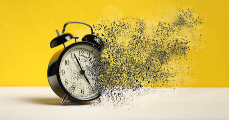 Alarm clock dissolving on white wooden table against yellow background, banner. Time is running out