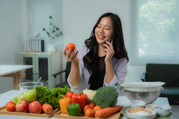 Asian nutritionist holding tomato and talking on phone with fresh vegetables lying on table in her...