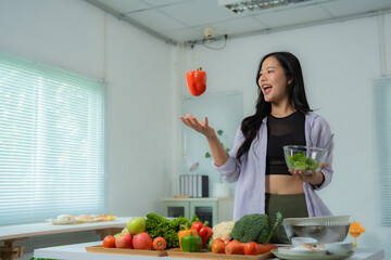 Young woman is juggling a bell pepper while holding a bowl of salad, surrounded by fresh produce in...