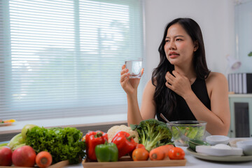 Young asian housewife having sore throat and drinking water while preparing healthy food in the...