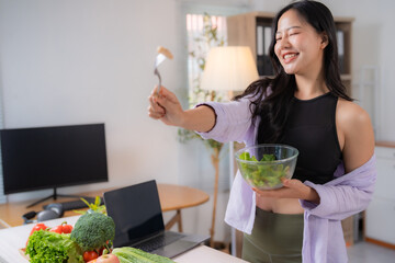 Smiling young woman in sportswear is holding a bowl of salad and a fork while having a video call on healthy eating habits from her home office