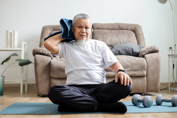 Fototapeta premium Elderly Asian man in sportswear wiping sweat on his face with towel during exercising at home