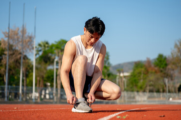 An asian man in white tank top is crouching while tying his sneaker shoelace on the racetrack.