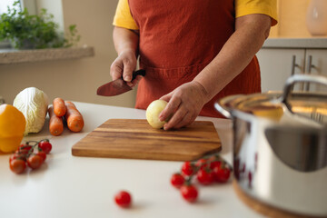 A woman in a yellow shirt and apron is peeling an onion in the kitchen. She is preparing ingredients for a meal while surrounded by fresh vegetables like carrots, tomatoes, and bell peppers