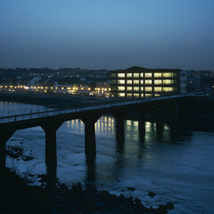Fototapeta premium Illuminated riverside building at dusk, city lights reflecting on water, bridge in foreground, urban landscape, perfect for real estate or travel websites