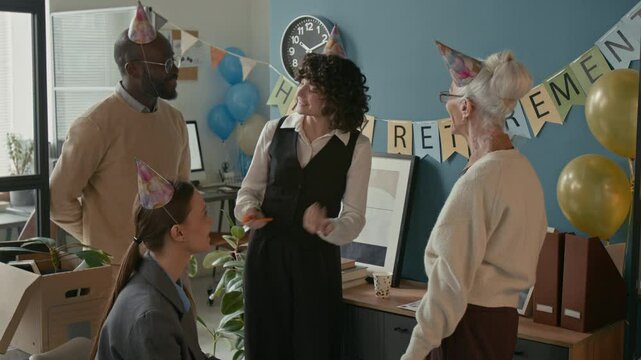 Young multiethnic coworkers presenting wrapped gift to smiling senior woman as part of her send-off celebration with balloons and decorations during joyful office retirement party