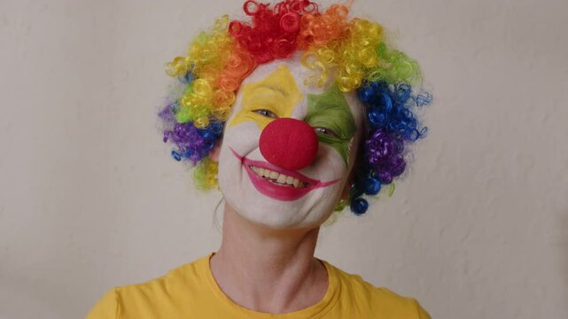 Portrait of clown in rainbow wig and red nose smiles and laughs looking at camera.
