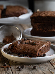 A slice of chocolate brownie garnished with cocoa and sprinkles. A stack of brownies in the background.
