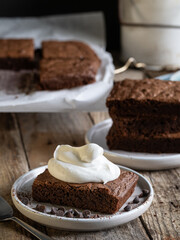 A slice of chocolate brownie with whipped cream. A stack of brownies in the background. A rustic-style table.