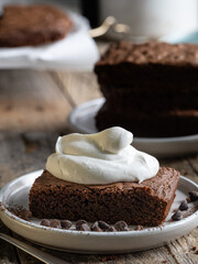 A slice of chocolate brownie with whipped cream. A stack of brownies in the background. A rustic-style table.