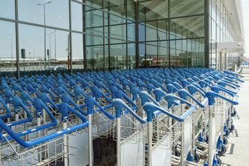 Neatly arranged blue airport trolleys line up outside a modern terminal reflecting in glass panels. Ready for passenger use, they promise convenience for travelers at the airport entrance.
