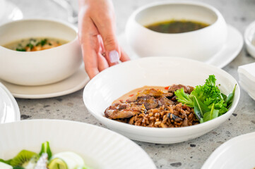 A hand reaching for a bowl of savory rice topped with tender meat and fresh greens, surrounded by other dishes, showcasing a flavorful and inviting dining experience.