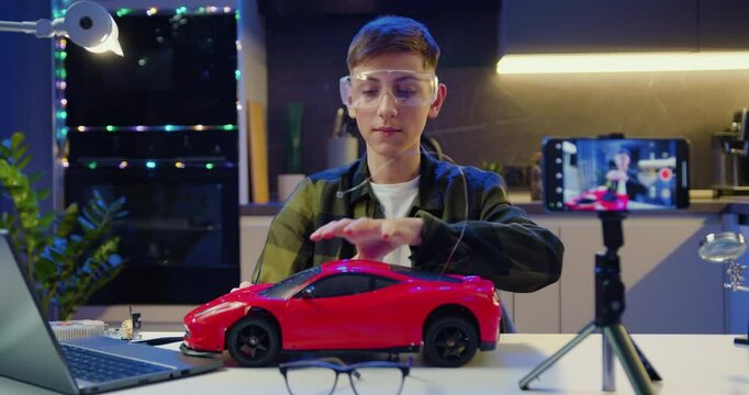 Teenage boy vlogger is recording video using smart phone about assembling and testing electric circuit building and programming car in evening at home.