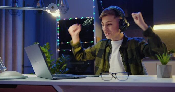 Teenage boy sits at desk using laptop reading e-mails online, gets good news, positive exam results and celebrates victory in evening at home.