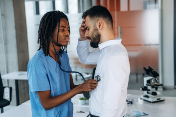 Doctor is using stethoscope, listening the heart of patient. Two men are in the office