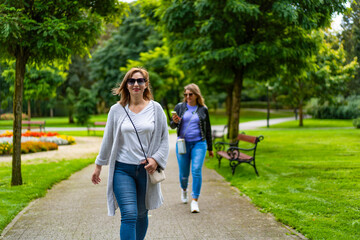 Obraz premium Two beautiful mature women walking separately, one goes in front, other in back, in city park. Front view