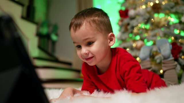 Beautiful smiling toddler in red t-shirt lies on white fluffy plaid. Joyful baby looks at tablet with interest. Close up. Christmas tree at backdrop.
