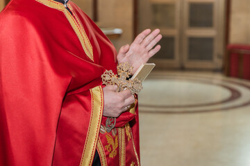 Orthodox priest in a red and gold robe, holding a golden cross and making a blessing gesture during...