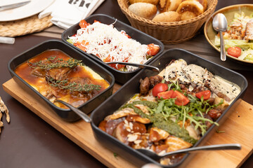 Delicious variety of main courses served in black dishes: hearty meat stew, Shopska salad with grated cheese, and sliced meat with potatoes, greens, and sauce.