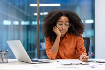 A woman with glasses sits at her desk, looking concerned while reviewing documents and a laptop. Her expression suggests she is stressed.