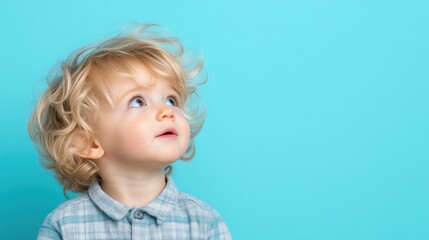 Adorable toddler gazing upward against a vibrant backdrop.  A sweet, light-skinned baby with curly blond hair looks thoughtfully upward, against a solid teal background.
