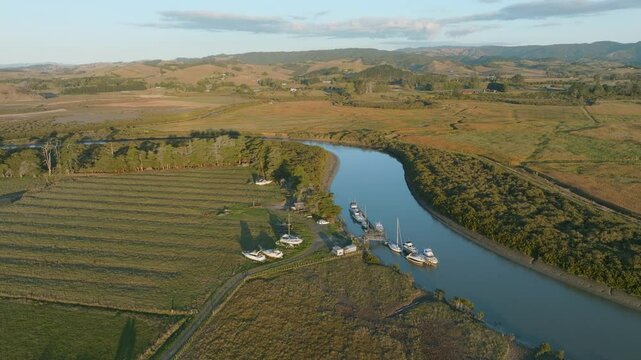 Aerial view of boats docked at a marina along a river in a rural landscape. The boats are moored to a wooden pier, and the surrounding area is a mix of farmland and natural vegetation. CLEVEDON, NZ