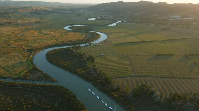 Aerial view of boats moored in a river, surrounded by lush greenery and farmland, bathed in the warm glow of the setting sun in New Zealand. CLEVEDON, NZ