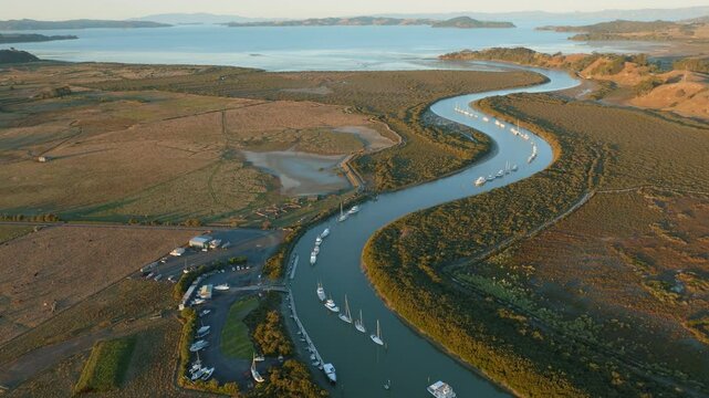 Aerial view of boats moored in a river, surrounded by lush greenery and mudflats, leading to the ocean in New Zealand. The scene captures the serene beauty of coastal life. CLEVEDON, NZ