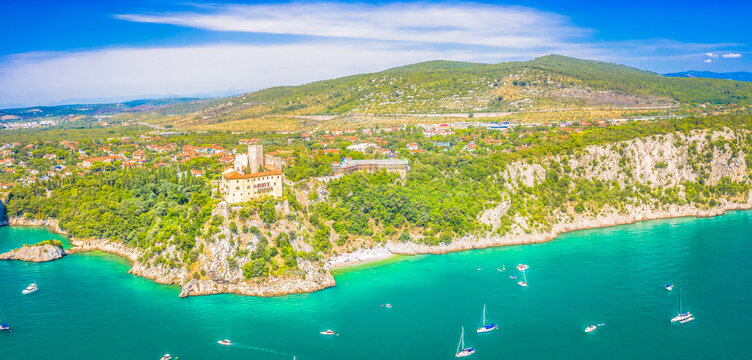 A wide panoramic view of the Adriatic coastline near Trieste, Italy, with the majestic landmark perched on a cliff, surrounded by lush greenery and overlooking the blue sea in a spectacular setting