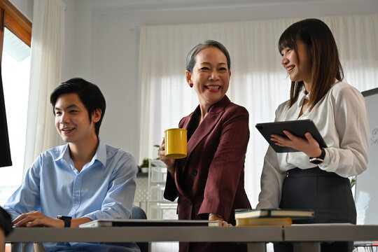Happy team of diverse professionals collaborating in a modern office. A senior woman holding a coffee mug chats with her colleagues using a desktop computer and tablet