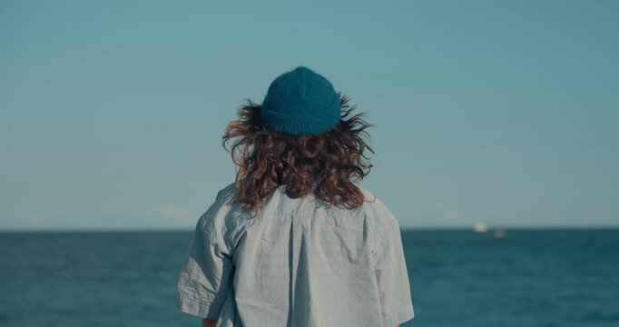 Authentic young woman in blue themed outfit, striped t-shirt, hand knitted blue wool beanie and prescription glasses and curly hair looks at ocean and then directly at camera. Candid image of youth