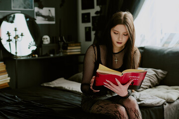 Medium shot of brilliant goth teen girl sitting on bed and enjoying reading book full of horror and...