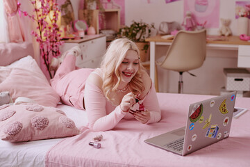 Smiling blonde teen girl lying on bed, polishing nails while enjoying process of watching videos via laptop