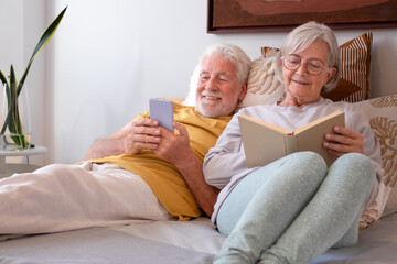 Portrait of serene senior couple relaxing together in bed, woman reading book, man using phone
