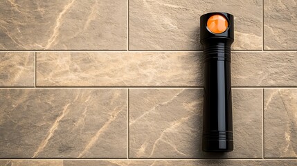 A sleek black flashlight with an orange button resting on textured stone tiles in a well-lit room