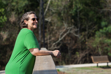A woman in a green shirt is standing on a wooden fence, smiling and looking out at the trees