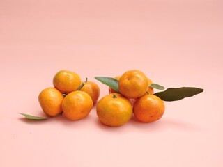 Vibrant santang or mandarin orange fruits with their characteristic textured skin, accompanied by natural green leaves, presented on a pastel pink backdrop. Illustrates freshness and natural produce.