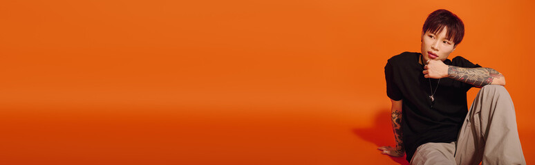 A fashionable young asian man leans casually against a vivid orange wall, showcasing modern style.