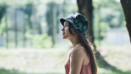 Young woman relaxing in green park, sporting cannabis themed bucket hat, radiating laid back summer...