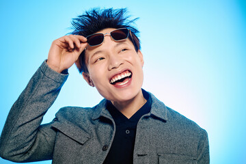 A young man with a trendy outfit joyfully adjusts his sunglasses while beaming a bright smile.