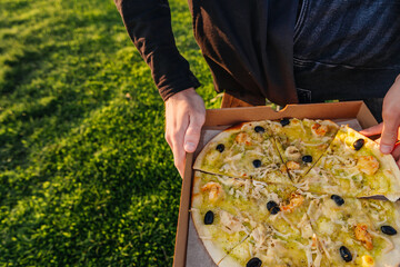 green pizza with seafood in a box in his hands on a background of green grass, the concept of a snack in nature