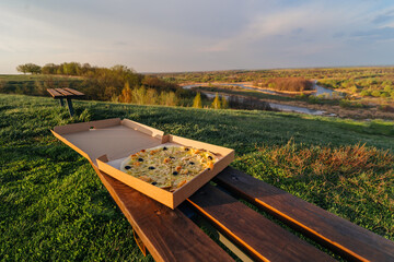 a pizza in a box stands on a bench on a hill with a beautiful view of the river, the concept of food in travel