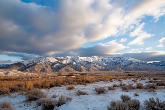 A breathtaking view of a snow-covered mountain range under a cloudy sky, showcasing shadows across the peaks and expansive open terrain in the foreground